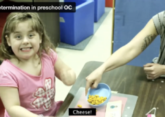 Screen grab from the Self-determination in Preschool video showing a young female student smiling at the camera while her teacher, sitting close by, shares some goldfish crackers with her.