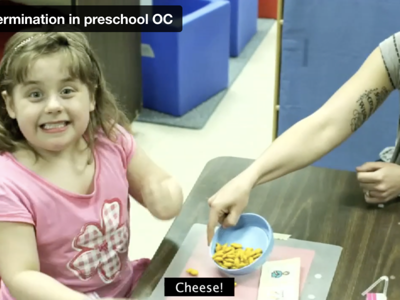 Screen grab from the Self-determination in Preschool video showing a young female student smiling at the camera while her teacher, sitting close by, shares some goldfish crackers with her.