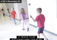 Young elementary school students walk through the hallway. One boy wearing a red shirt and black shorts is waving.