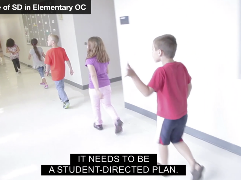 Young elementary school students walk through the hallway. One boy wearing a red shirt and black shorts is waving.