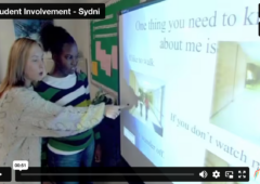 Screenshot of video showing Sydni and her teacher at a SmartBoard, pointing to her IEP presentation.