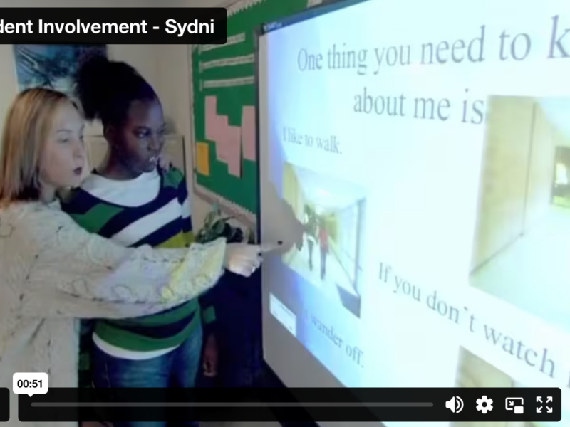 Screenshot of video showing Sydni and her teacher at a SmartBoard, pointing to her IEP presentation.
