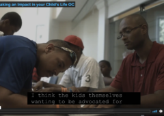 Screen capture of video screen showing two men at a table. One is filling out a form and the other is assisting.