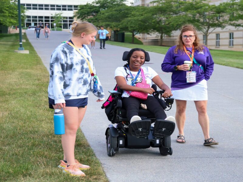 Three young women, one in a wheelchair, make their way down a long sidewalk on a college campus.