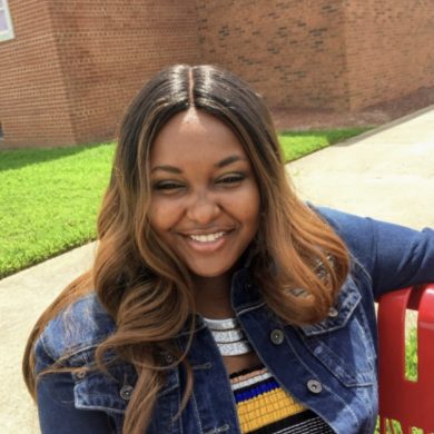 Headshot of youth leader, Traleya, female with dark brown hair with highlights, wearing a striped shirt and jean jacket, sitting on a bench outdoors