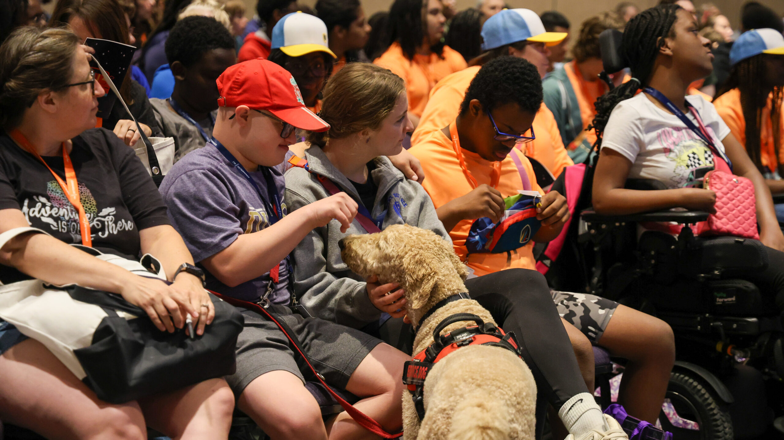 Group shot of participants at the 2024 Youth & Family Summit, sitting in the main ballroom before a session begins. In front a young man with a red hat sits next to a member of the support staff while they pet his service dog.