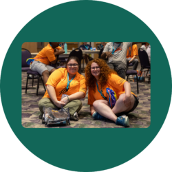 Youth Leaders Kyla and Liat sitting on the floor of the ballroom at the Youth & Family Summit wearing bright orange Youth Leader t-shirts.