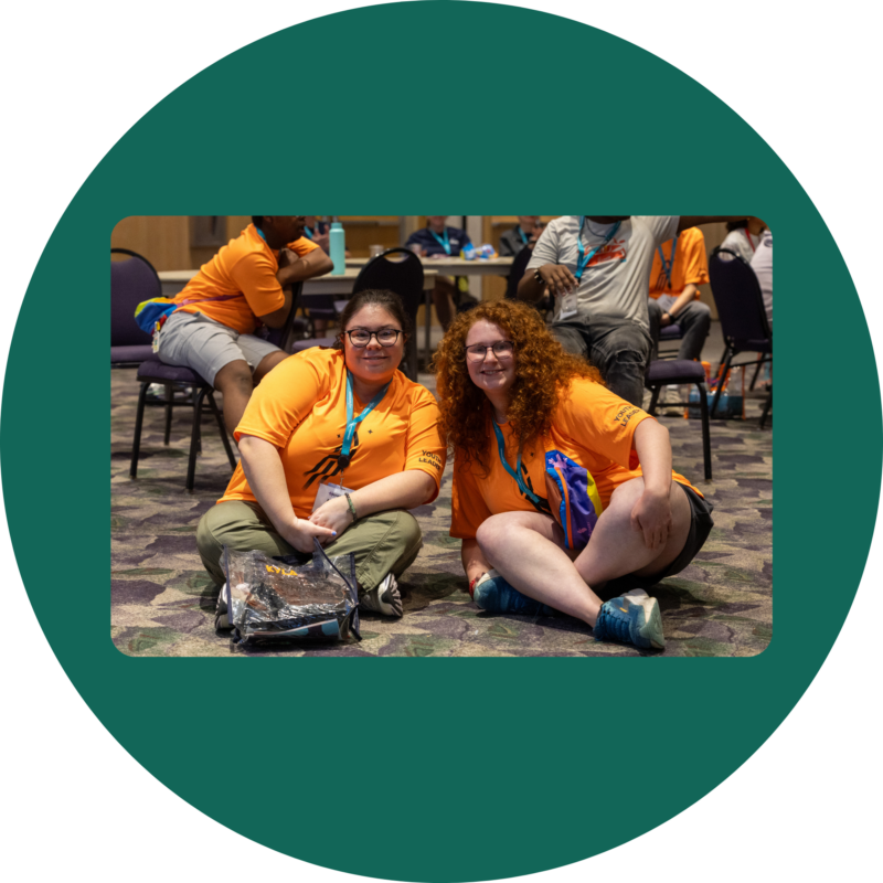 Youth Leaders Kyla and Liat sitting on the floor of the ballroom at the Youth & Family Summit wearing bright orange Youth Leader t-shirts.