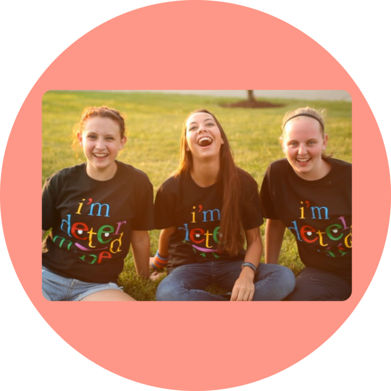 Three I'm Determined Youth Leaders sit on the grass, smiling, wearing a black t-shirt with the I'm Determined logo on the front.